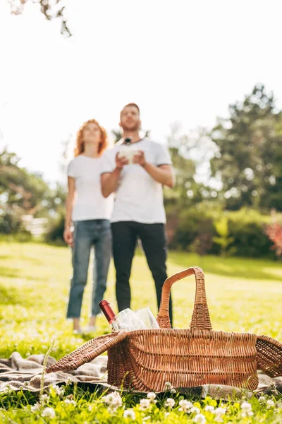 Vista de cerca de la cesta de picnic y pareja jugando con el dron detrás en el parque - foto de stock