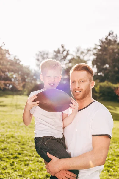 Happy father and son holding rugby ball and smiling at camera in park — Stock Photo
