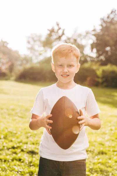 Adorable child holding rugby ball and smiling at camera in park — Stock Photo