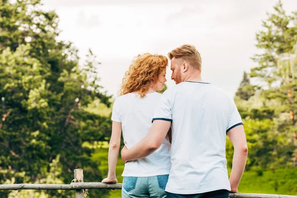 Vista trasera de pareja pelirroja abrazándose y sonriéndose mientras están de pie juntos en el parque - foto de stock