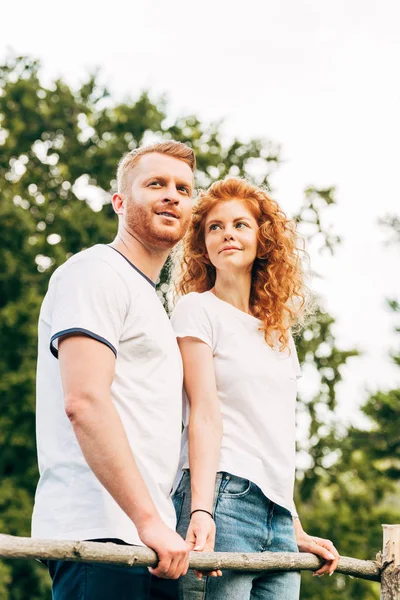 Vista basso angolo di felice rossa coppia in piedi insieme e guardando lontano al parco — Foto stock