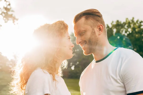 Vista lateral de la feliz pareja pelirroja sonriéndose mientras están de pie en el parque al atardecer - foto de stock