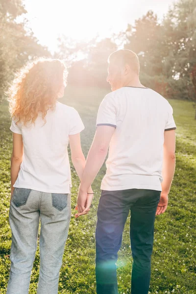 Vista trasera de joven pelirroja pareja cogida de la mano y caminando juntos en el parque - foto de stock