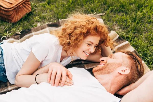 Feliz joven pelirroja pareja acostado en cuadros y sonriendo el uno al otro en el picnic - foto de stock