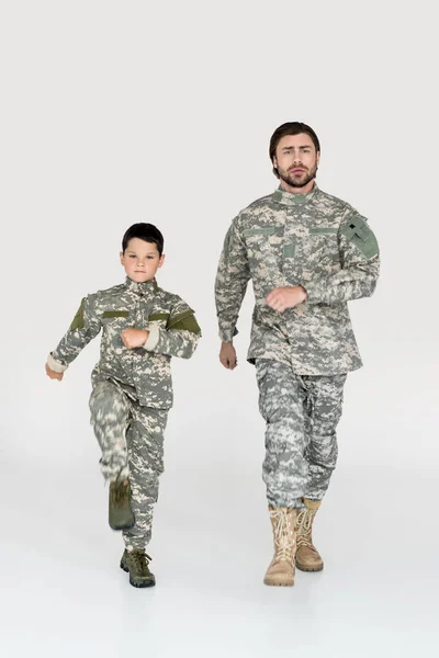 Padre e hijo en uniformes militares marchando y mirando a la cámara sobre fondo gris - foto de stock