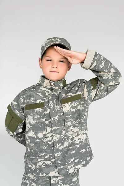 Портрет маленького мальчика в военной форме, салютующего на сером фоне — стоковое фото