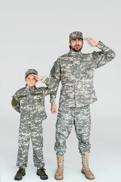 Father and son in military uniforms saluting and looking at camera on grey background — Stock Photo