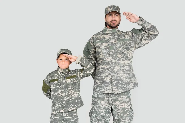 Retrato de soldado e filho confiantes em uniformes militares saudando isolados em cinza — Fotografia de Stock