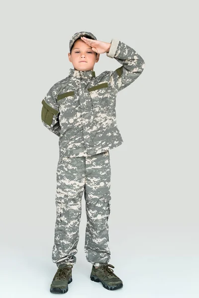 Kleines Kind in Militäruniform blickt in die Kamera und salutiert auf grauem Hintergrund — Stockfoto