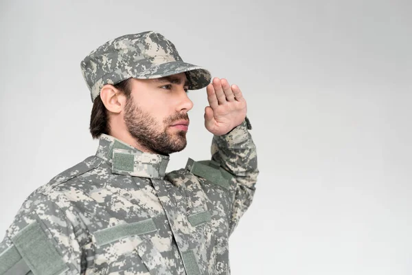 Vista lateral de soldado barbudo confiado en uniforme militar saludando sobre fondo gris - foto de stock