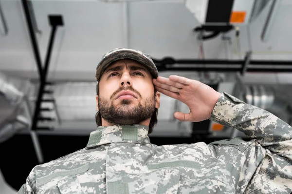 Vista de ángulo bajo de soldado confiado en uniforme militar saludando con fondo borroso - foto de stock