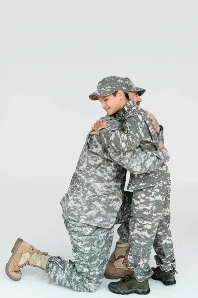 Side view of father and son in military uniforms hugging each other on grey background — Stock Photo