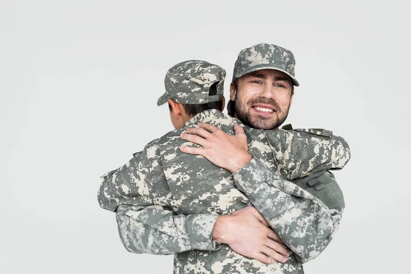 Partial view of smiling soldier and son in military uniforms hugging each other isolated on grey — Stock Photo