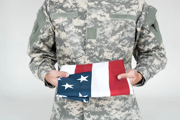 Vista parcial del soldado en uniforme militar con bandera americana en las manos sobre fondo gris — Stock Photo