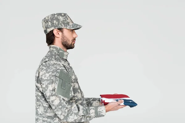 Vista lateral do soldado masculino em uniforme militar com bandeira americana em mãos em pano de fundo cinza — Fotografia de Stock