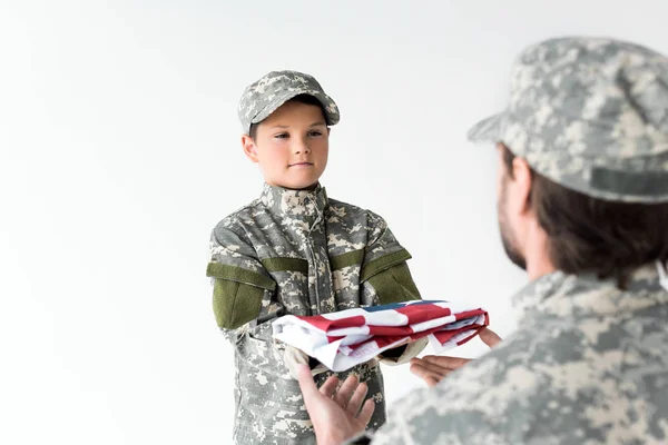 Частковий вигляд маленького хлопчика в камуфляжному одязі, що дає складений американський прапор солдату на сірому фоні — стокове фото