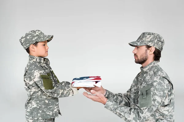 Vista lateral del niño pequeño en ropa de camuflaje dando bandera americana plegada al soldado sobre fondo gris - foto de stock