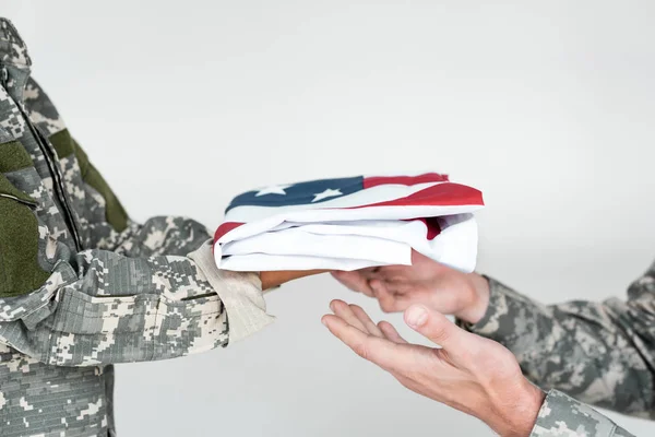 Cropped shot of kid in camouflage clothing giving folded american flag to soldier on grey background — Stock Photo