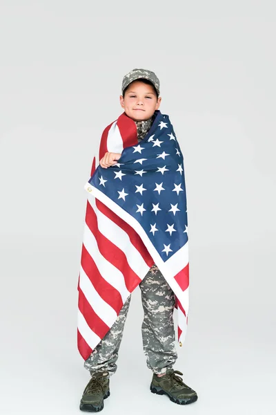 Niño en uniforme militar cubierto con bandera americana mirando a la cámara sobre fondo gris - foto de stock