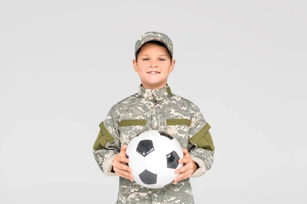 Retrato de niño sonriente en uniforme militar con pelota de fútbol en manos aisladas en gris - foto de stock