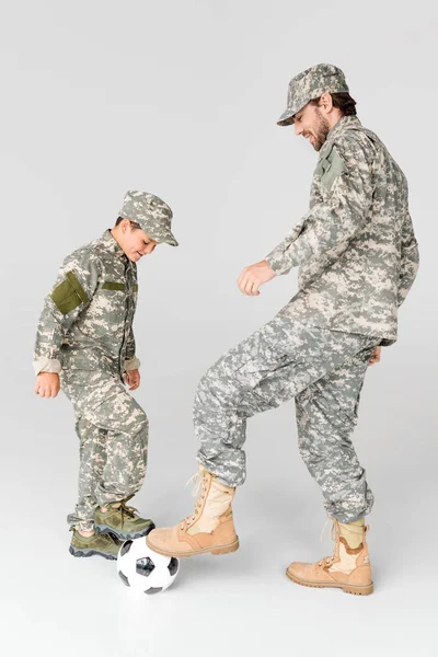 Вид сбоку отца и сына в военной форме, играющих в футбол на сером фоне — стоковое фото