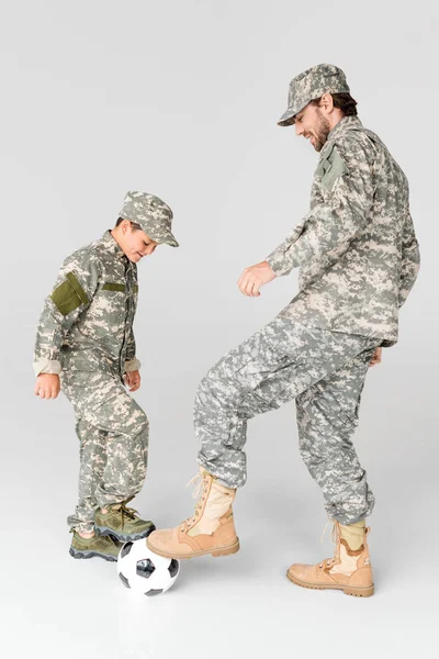 Vista laterale di padre e figlio in uniforme militare che giocano a calcio su sfondo grigio — Foto stock