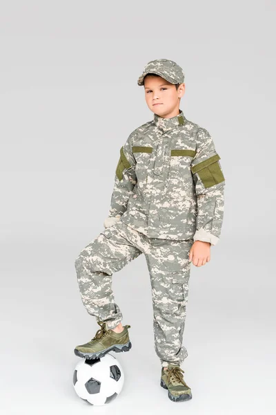 Kid in military uniform with soccer ball looking at camera isolated on grey — Stock Photo