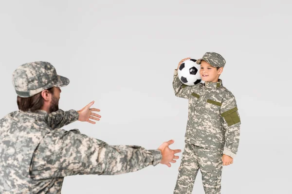 Soldier with outstretched arms and smiling kid in camouflage clothing with soccer ball isolated on grey — Stock Photo