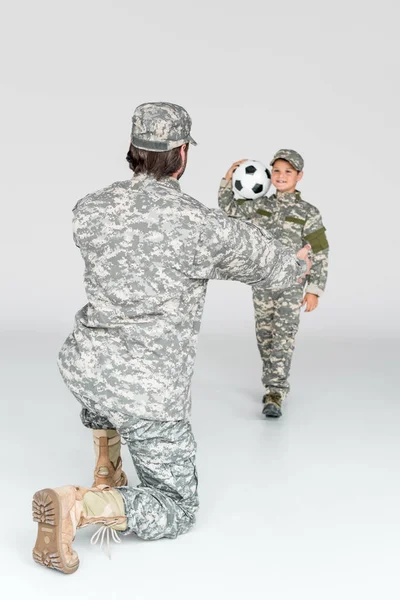 Vista parcial de soldado con los brazos extendidos y niño sonriente en ropa de camuflaje con pelota de fútbol sobre fondo gris - foto de stock