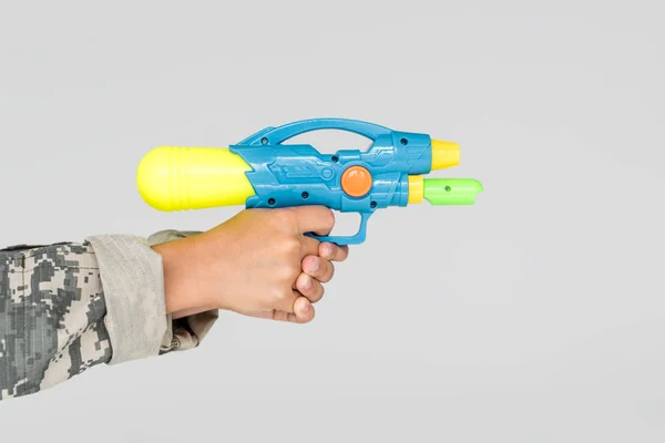 Vista parcial de niño en ropa de camuflaje con pistola de agua de juguete en manos aisladas en gris - foto de stock