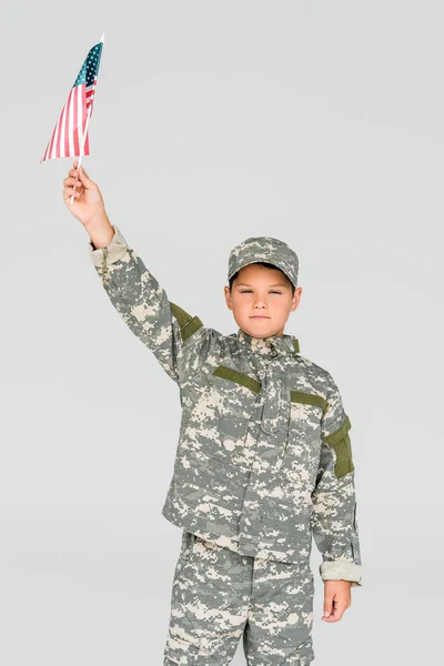 Retrato de menino em roupas de camuflagem segurando bandeira americana na mão isolado em cinza — Fotografia de Stock