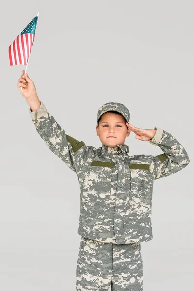 Portrait of boy in camouflage clothing saluting while holding american flagpole in hand isolated on grey — Stock Photo