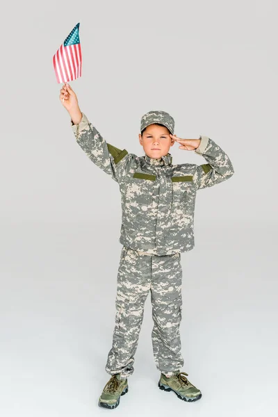 Menino em camuflagem roupas saudando enquanto segurando bandeira americana na mão isolado em cinza — Fotografia de Stock