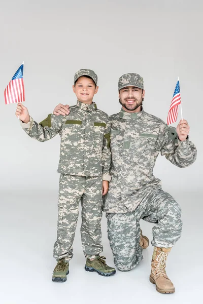 Улыбающиеся отец и сын в камуфляжной одежде с американскими флагштоками в руках на сером фоне — стоковое фото