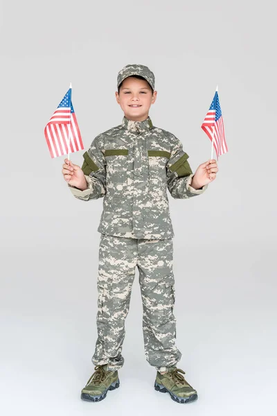 Enfant souriant en uniforme militaire avec des mâts américains dans les mains regardant la caméra sur fond gris — Photo de stock