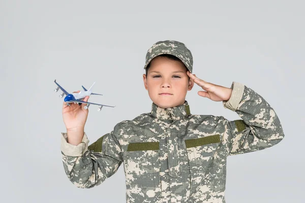 Retrato de niño pequeño en uniforme militar con avión de juguete en mano saludo aislado en gris - foto de stock