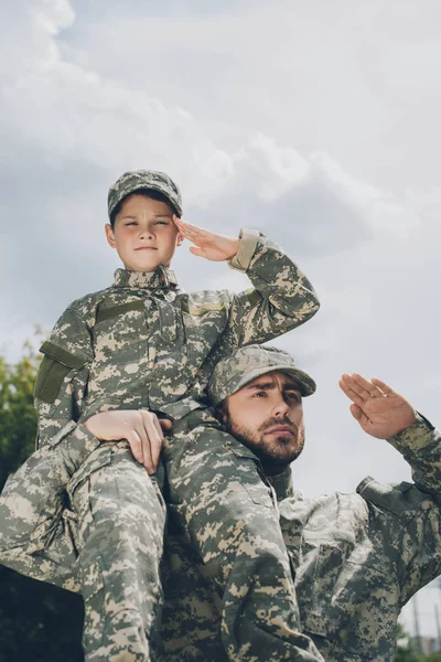 Vue à angle bas de la famille en uniforme militaire saluant avec ciel nuageux sur toile de fond — Photo de stock