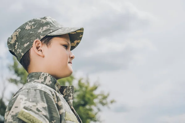 Vue latérale du petit garçon en tenue de camouflage avec ciel nuageux en toile de fond — Photo de stock