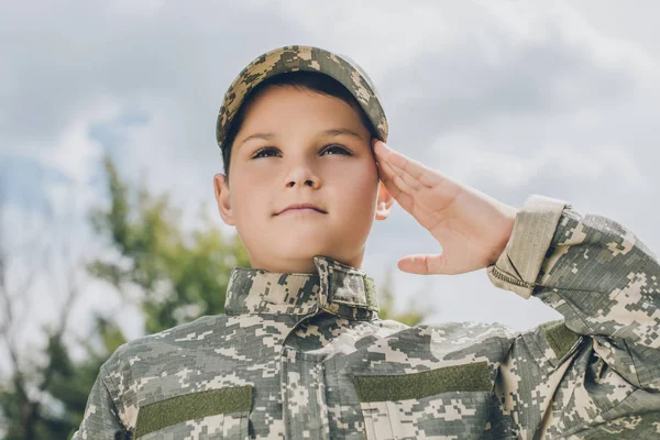 Portrait of little boy in camouflage clothing saluting with cloudy sky on backdrop — Stock Photo