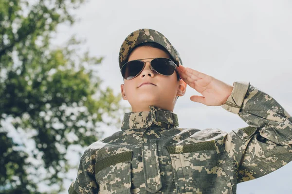 Retrato de niño pequeño en ropa de camuflaje y gafas de sol saludando con cielo nublado en el telón de fondo - foto de stock