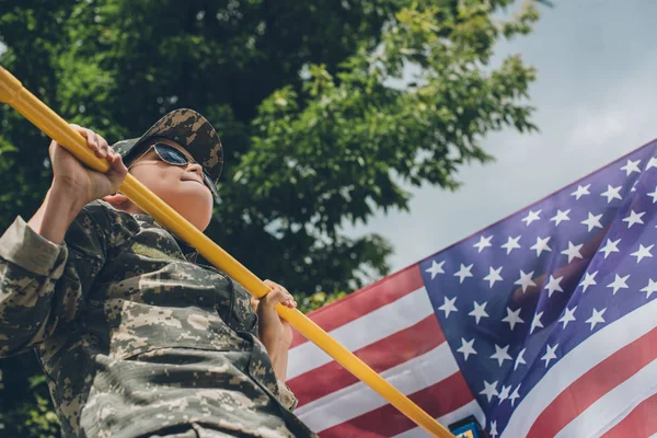 Low angle view of boy in sunglasses pulling himself up on crossbar with american flag on backdrop — Stock Photo