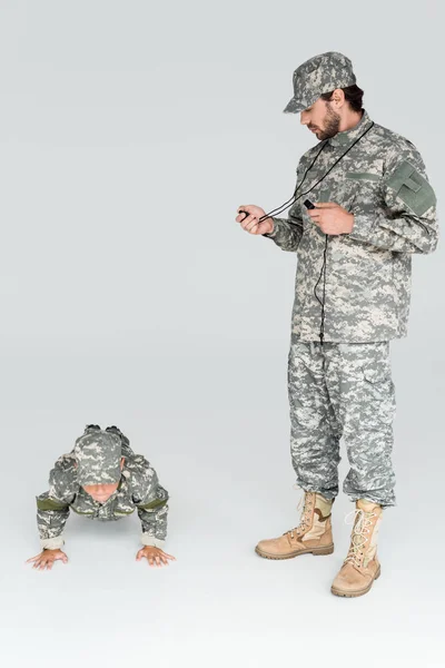 Soldado em uniforme militar com temporizador e assobio controlando o tempo enquanto filho fazendo flexões em fundo cinza — Fotografia de Stock