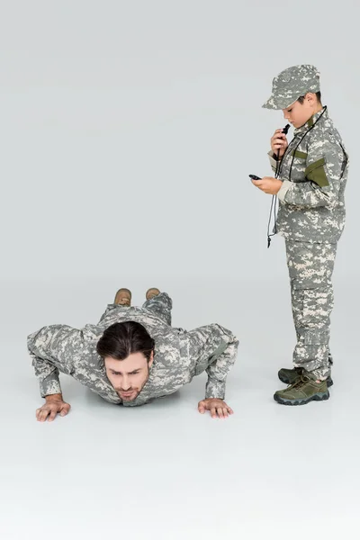 Menino em uniforme militar com temporizador e assobio controlando o tempo enquanto soldado fazendo flexões em fundo cinza — Fotografia de Stock