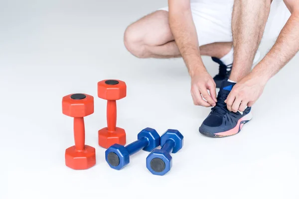 Vista parcial del hombre atando cordones cerca de pesas rojas y azules sobre fondo gris - foto de stock