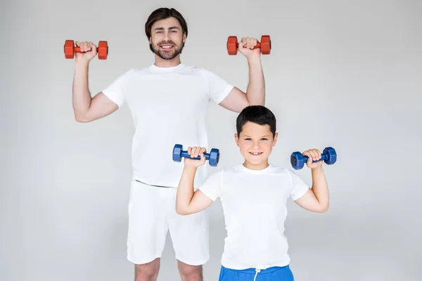 Retrato de pai e filho sorridentes em camisas brancas exercitando com halteres em fundo cinza — Fotografia de Stock