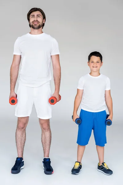 Padre e figlio sorridenti in camicie bianche che si esercitano con i manubri su sfondo grigio — Foto stock