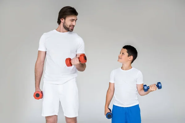 Smiling father and son in white shirts looking at each other while exercising with dumbbells on grey background — Stock Photo