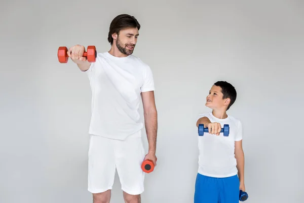 Smiling father and son in white shirts looking at each other while exercising with dumbbells on grey background — Stock Photo
