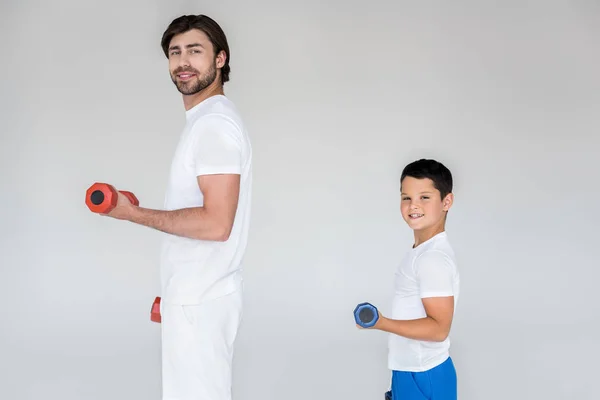 Vista lateral de pai e filho sorridentes em camisas brancas exercitando com halteres em fundo cinza — Fotografia de Stock