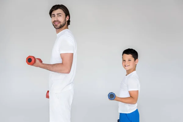 Side view of smiling father and son in white shirts exercising with dumbbells on grey background — Stock Photo